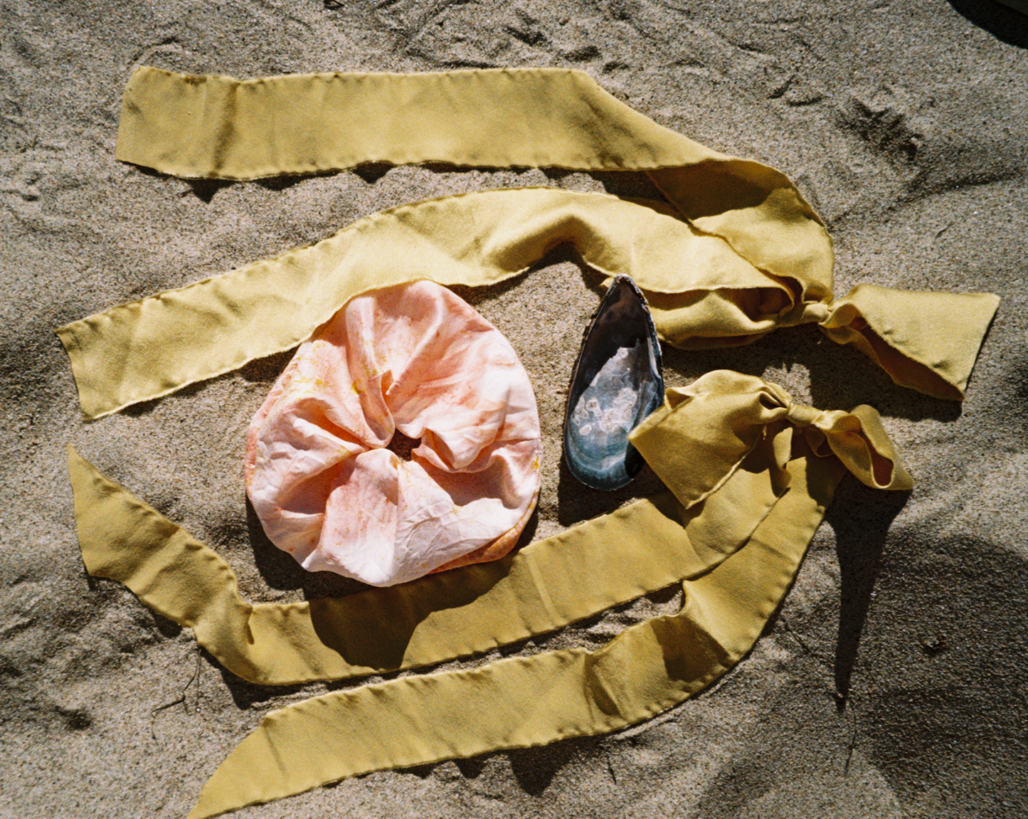 Two Silk green plant dyed bows are on the beach surround a mussel shell and a peach colored silk scrunchie
