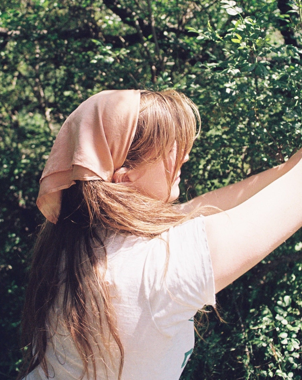 Women with long blonde hair is wearing a peach silk plant dyed bandana wrapped around her hair. She is surrounded by trees.