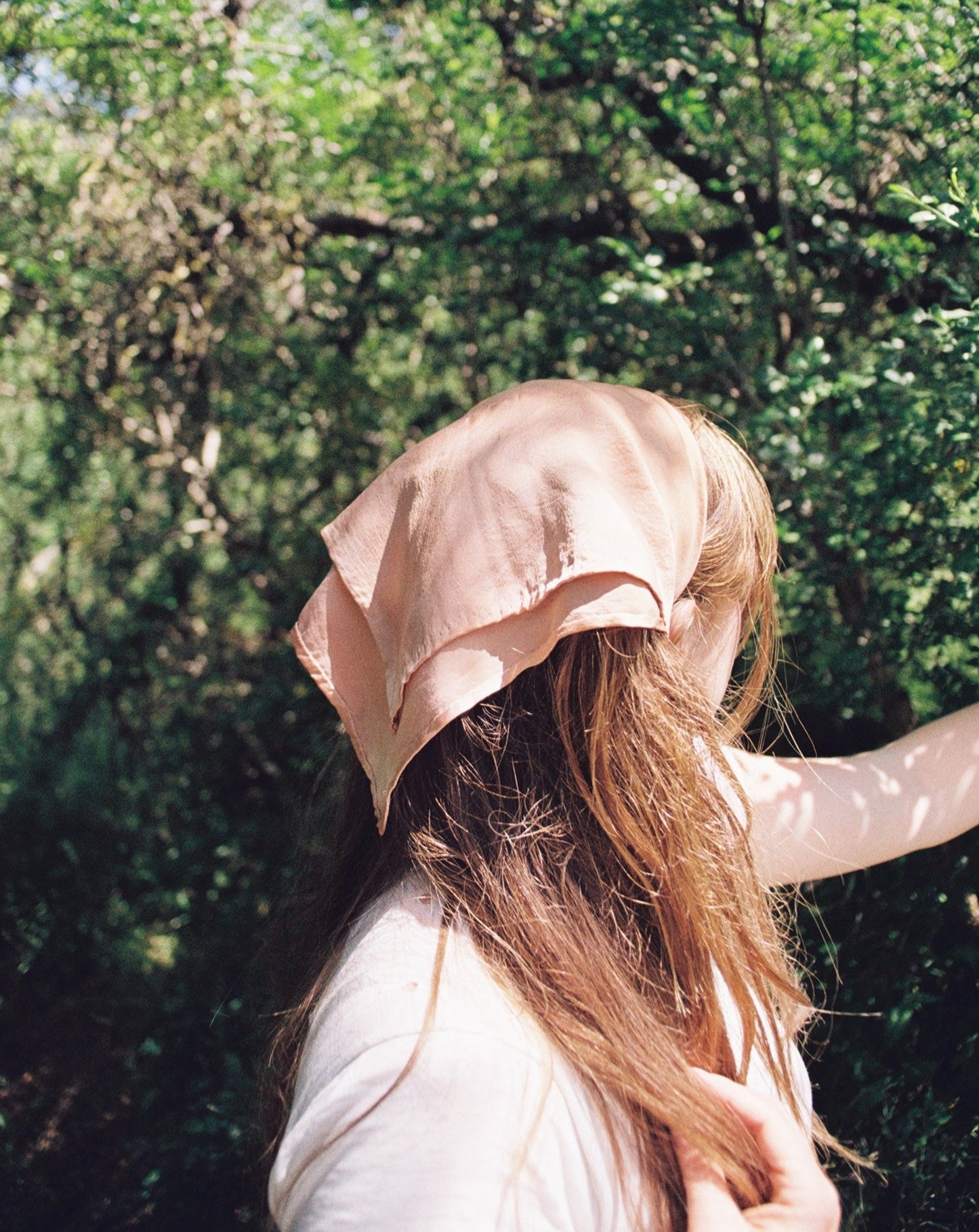 Women with long blonde hair is wearing a peach silk plant dyed bandana wrapped around her hair. She is surrounded by trees.