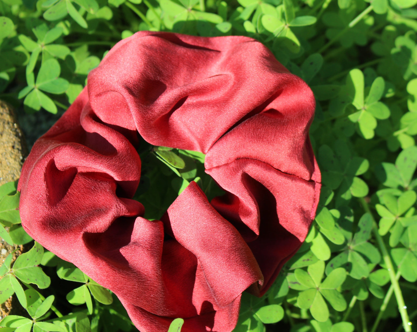 A ruby red silk scrunchie dyed with madder root to make a striking crimson red silk scrunchie, laying in a bed of clovers