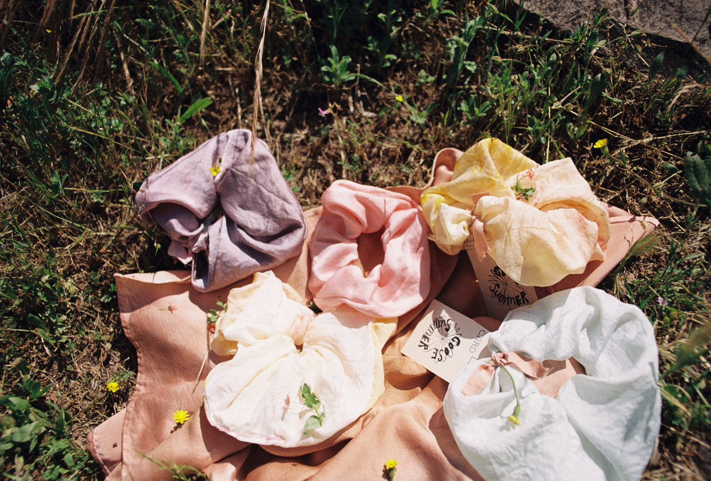 Five plant dyed silk scrunchie are laying in a field of grass. Hand made my Goose Summer Plant Dyes.Goose Summer is a small, sustainable plant dyeing business making naturally dyed silk scrunchies and other accessories in Los Angeles.