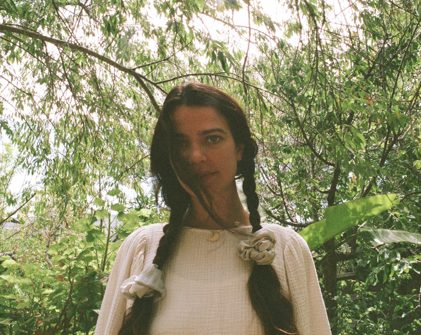 Woman wearing her hair in braids are tied with light blue naturally dyed silk scrunchies. She is wearing Goose Summer's Dawn silk scrunchies. She is surrounded by foliage.