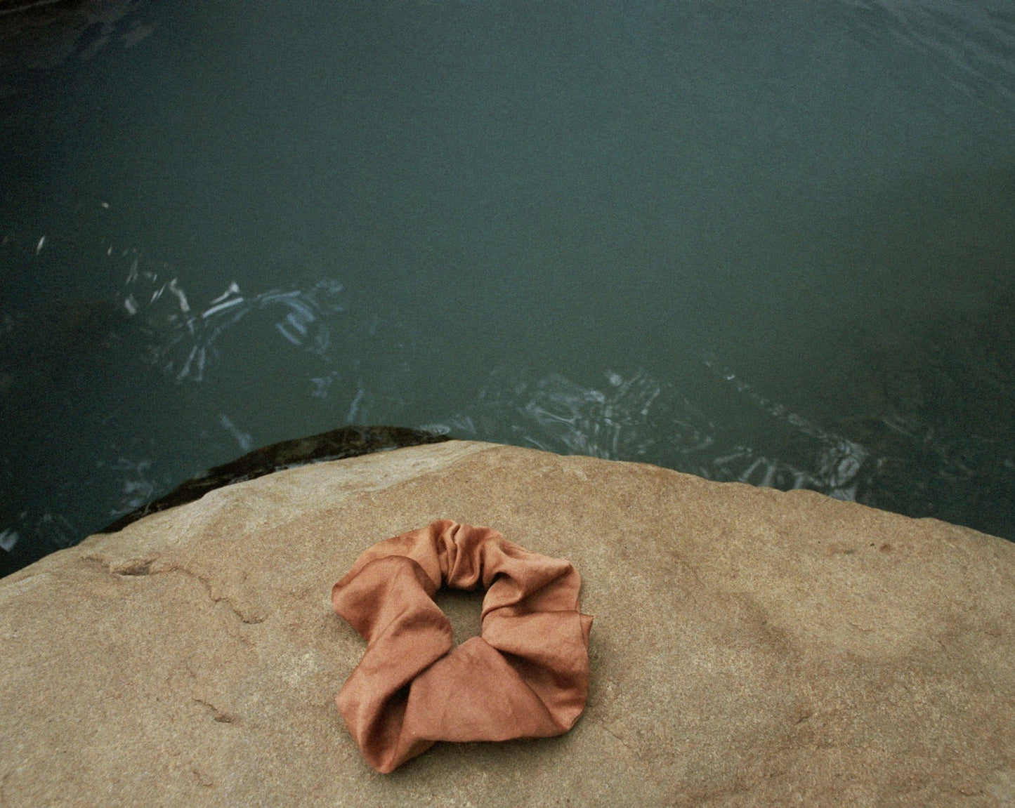 A brownish peach colored silk scrunchie is displayed on a rock. The Sienna Silk Scrunchie sits on a rock beside a hot spring. The Sienna Scrunchie is naturally dyed by Goose Summer with Cutch bark.