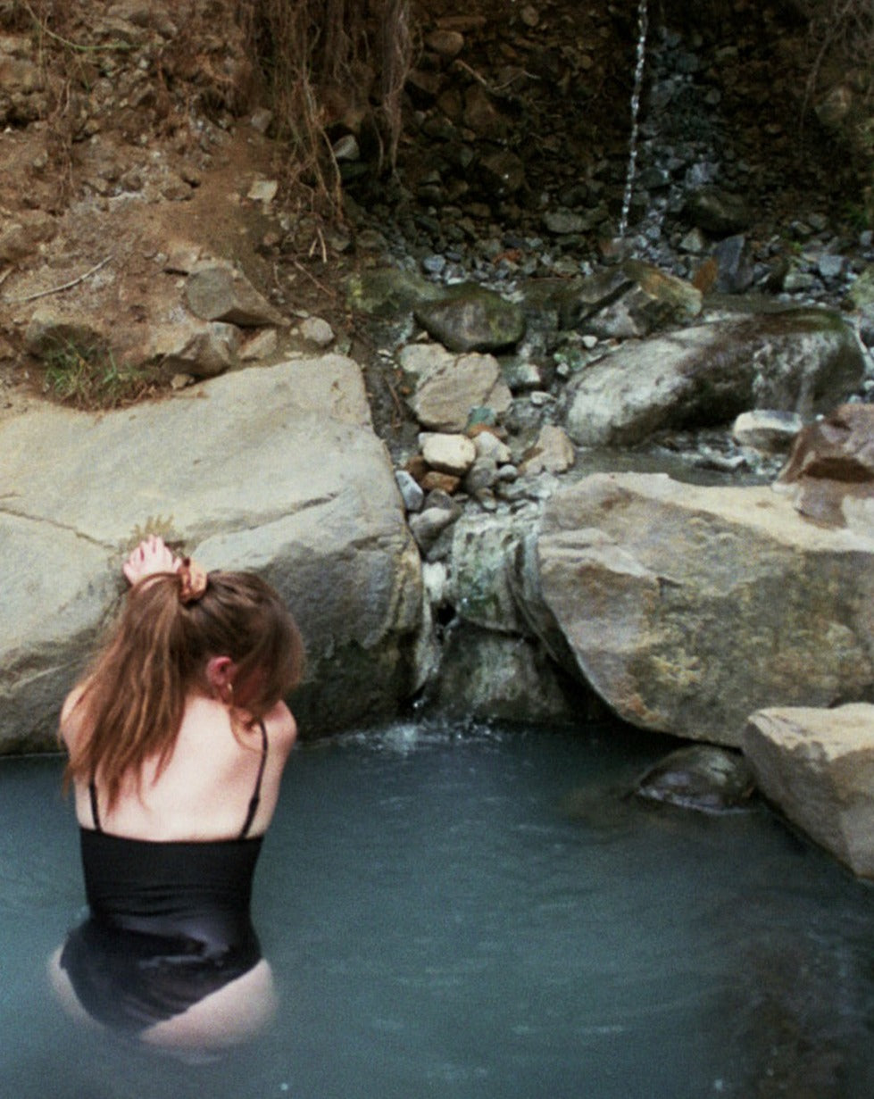 Woman leaning against a rock with her blonde hair tied in a Goose Summer naturally dyed silk scrunchie. She is wearing a brown silk scrunchie which is dyed with cutch bark.