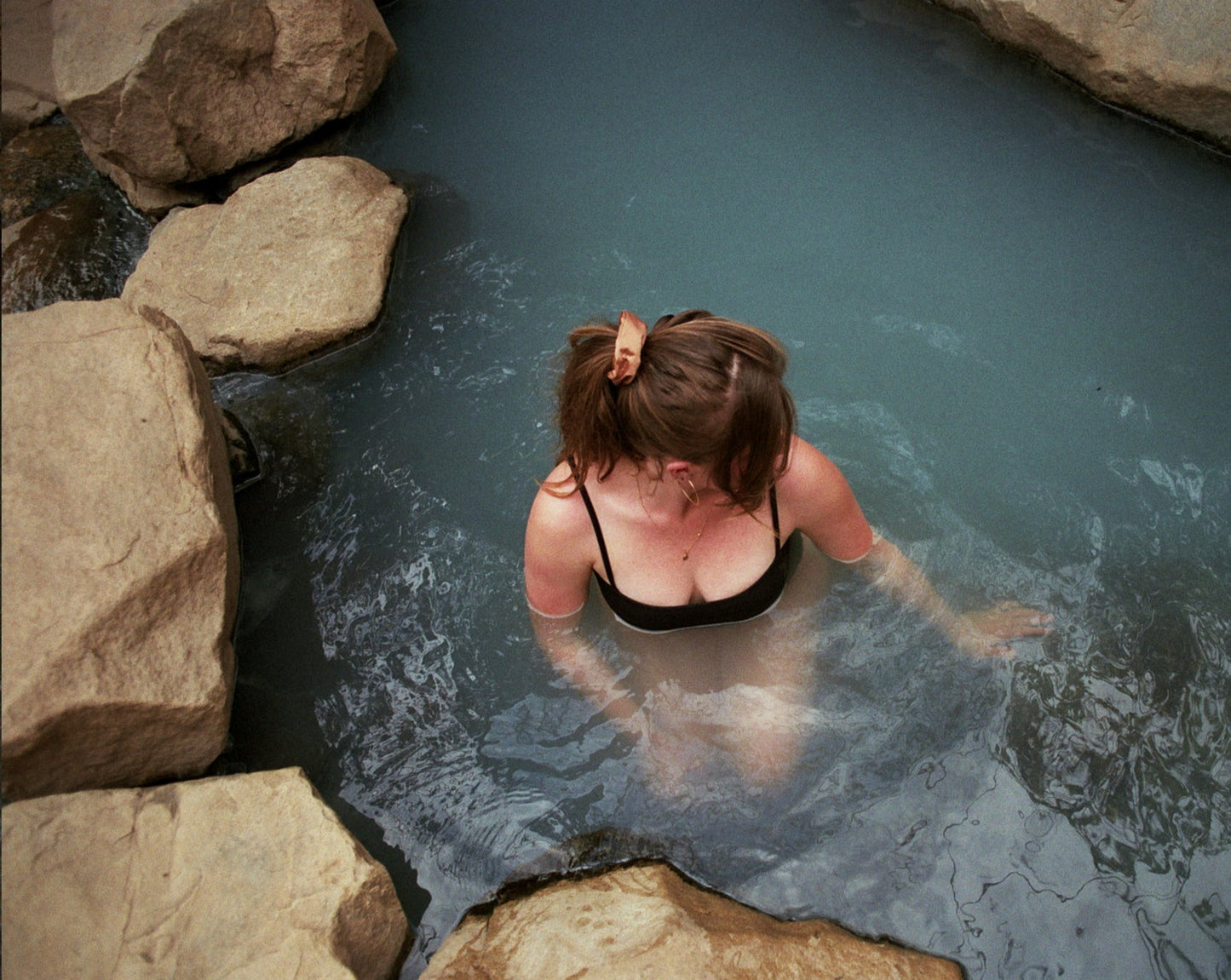 Woman swimming in a hot spring in a black bathing suit with her hair tied up in a ponytail with a Goose Summer naturally dyed brown silk scrunchie. She is wearing the Sienna Silk Scrunchie which is plant dyed with cutch bark.