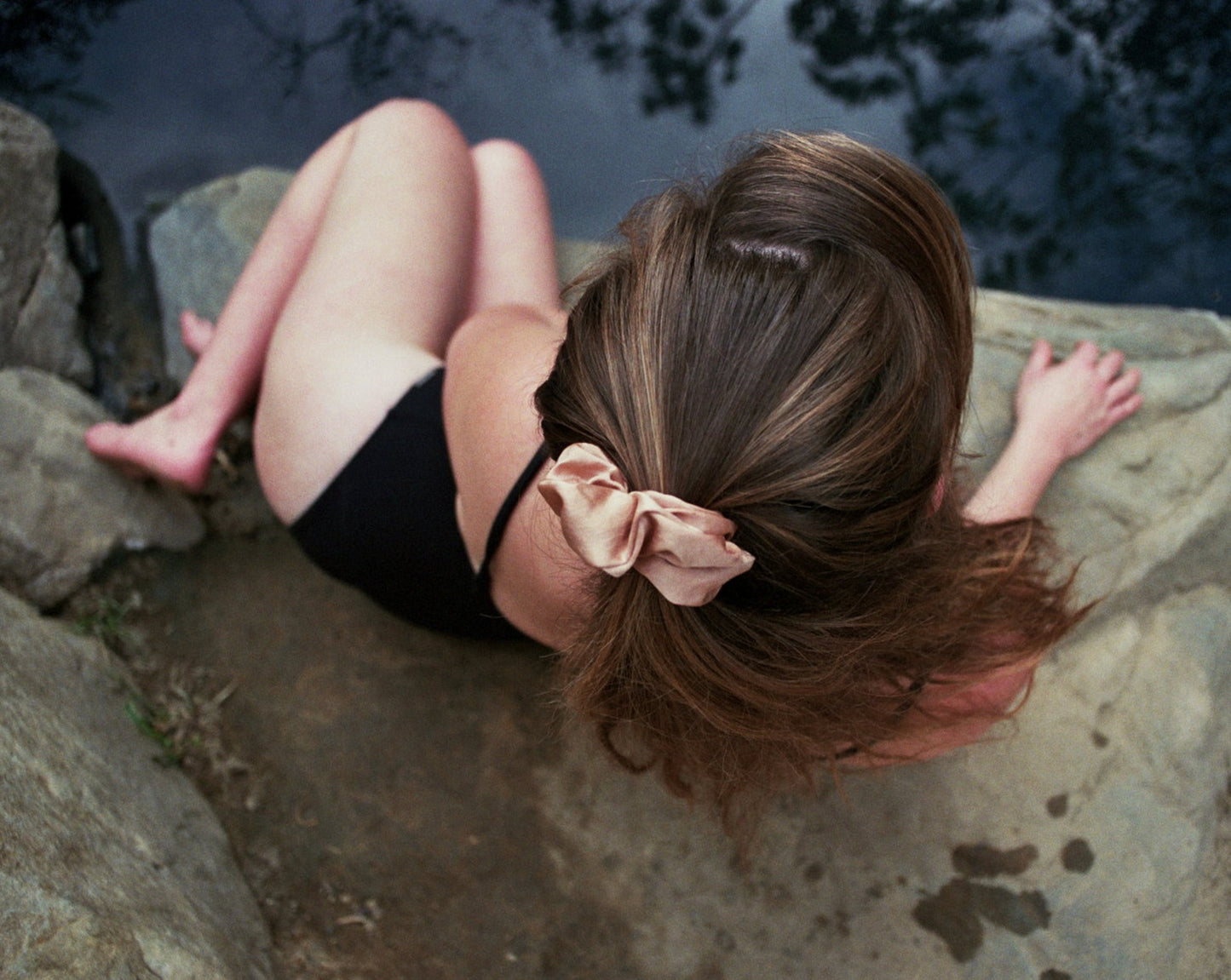 Woman sitting on a rock in a black bathing suit wearing the Goose Summer Apricot Scrunchie. This silk scrunchie is light pink, naturally dyed with cutch bark.