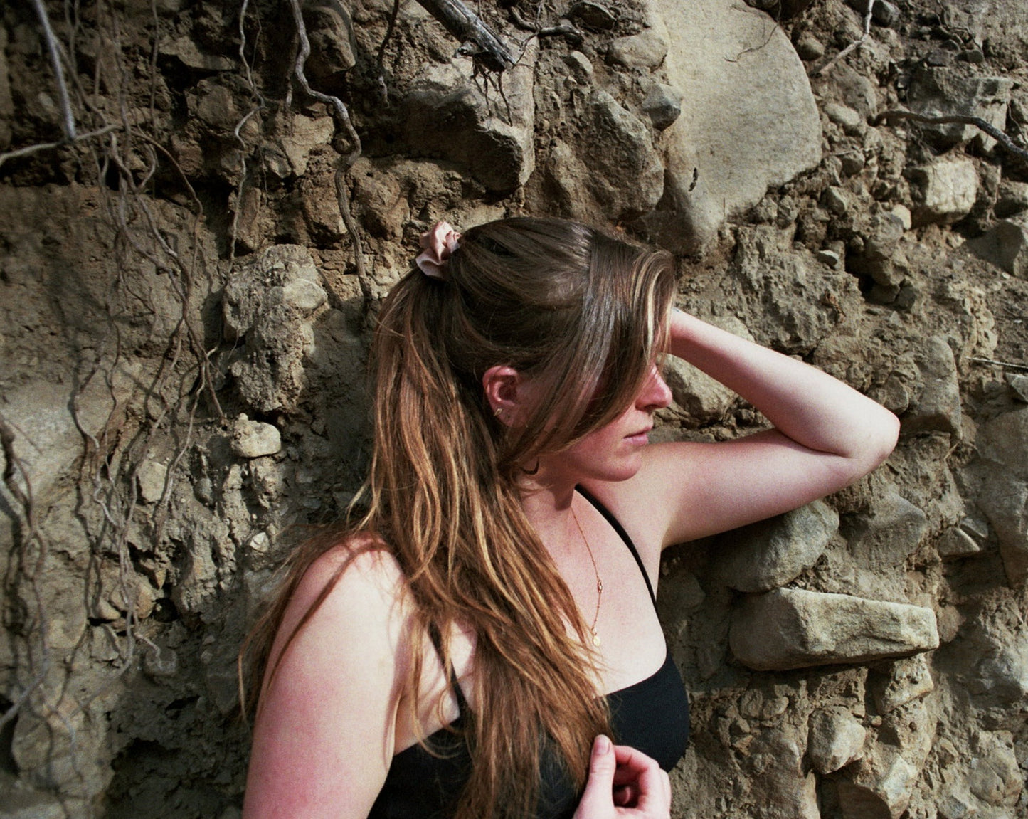 Woman posing against some rocks in nature with her hair tied in a ponytail with a light pink Goose Summer silk scrunchie. She is wearing the Apricot Scrunchie, which is naturally dyed with cutch bark.