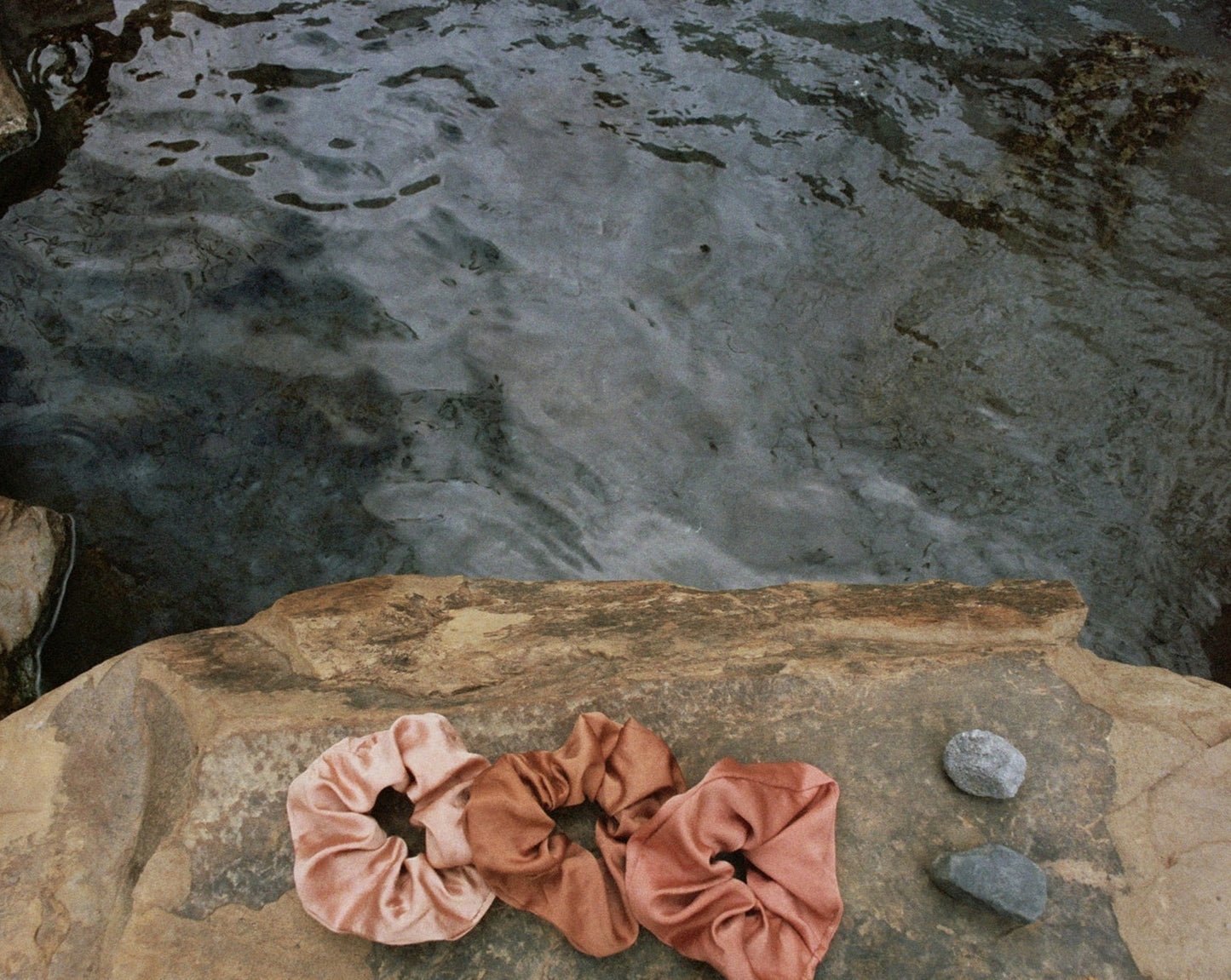 Three naturally dyed Goose Summer silk scrunchies are displayed on a rock in front of a hot spring. The colors of scrunchies from left the right are light pink, brown and rose pink silk scrunchies. Their names are Apricot scrunchie, Sienna Scrunchie, and Rose Scrunchie.