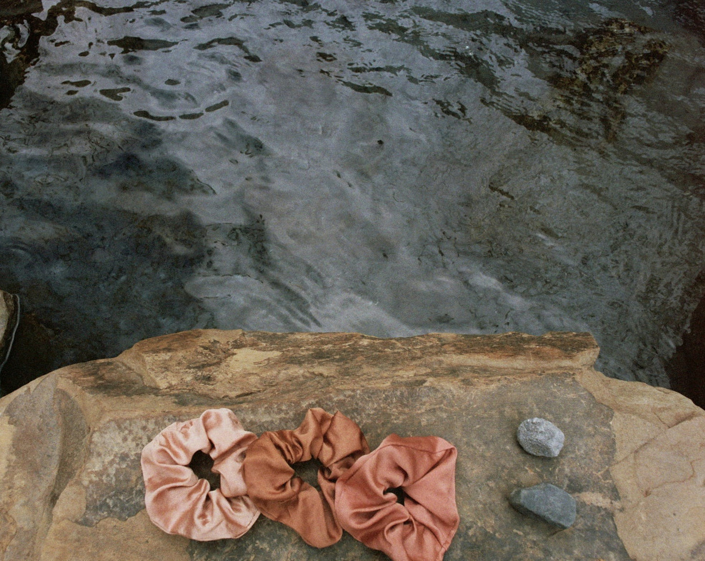 Three naturally dyed Goose Summer silk scrunchies are displayed on a rock in front of a hot spring. The colors of scrunchies from left the right are light pink, brown and rose pink silk scrunchies. Their names are Apricot scrunchie, Sienna Scrunchie, and Rose Scrunchie.