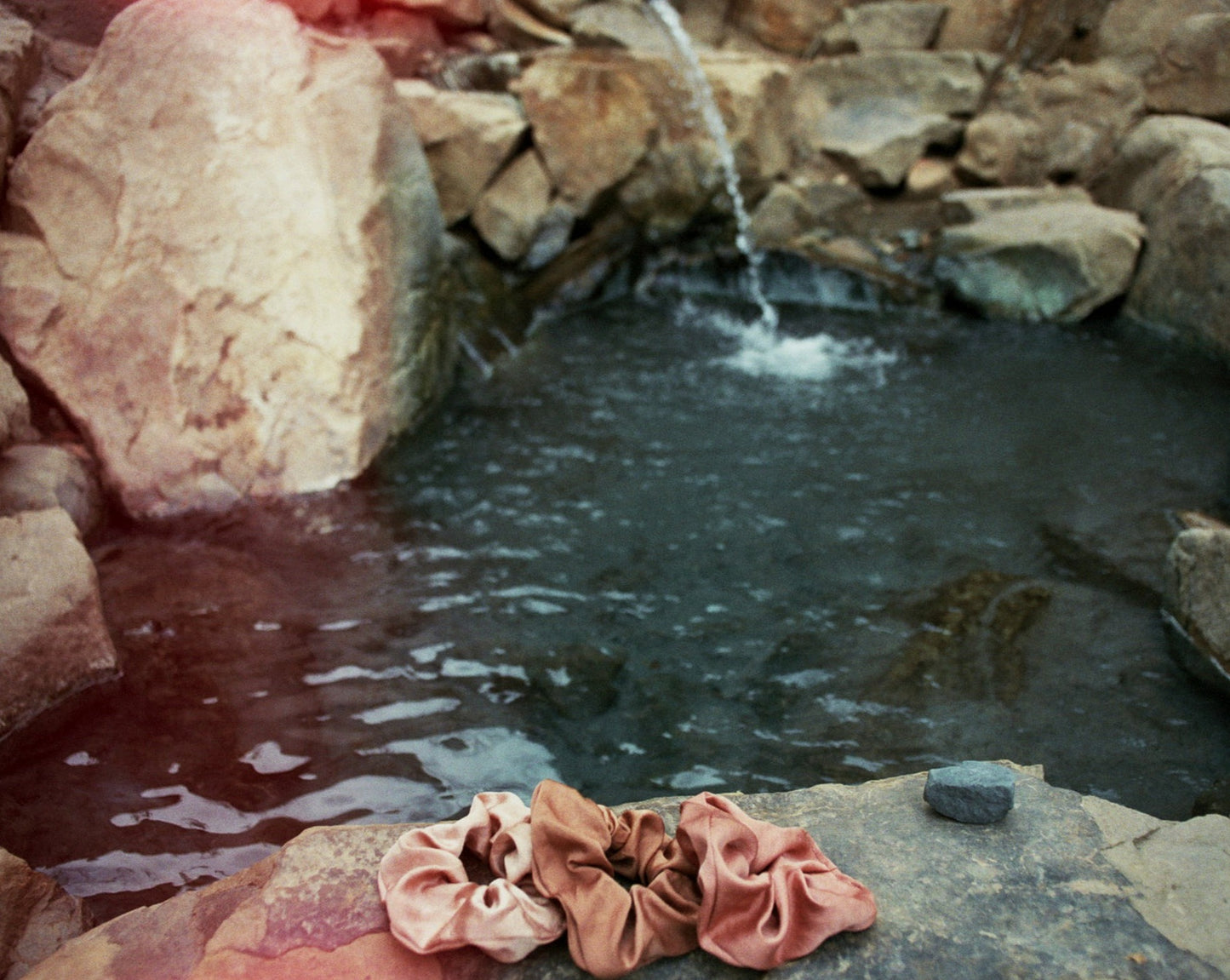 Three naturally dyed Goose Summer silk scrunchies are displayed on a rock in front of a hot spring. The colors of scrunchies from left the right are light pink, brown and rose pink silk scrunchies. Their names are Apricot scrunchie, Sienna Scrunchie, and Rose Scrunchie.