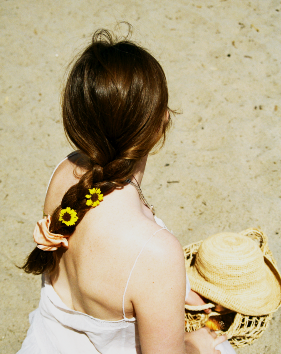 Woman sitting in the sand on the beach wearing her hair in one braid tied with a peach silk scrunchie. Peach is dyed naturally with madder root by Goose Summer. Madder Root is a plant dye that has been cultivated for hundreds of years as a natural textile dye. She has a peach scrunchie and yellow wild flowers in her hair.