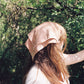 Women with long blonde hair is wearing a  peach silk plant dyed bandana wrapped around her hair. She is surrounded by trees. 