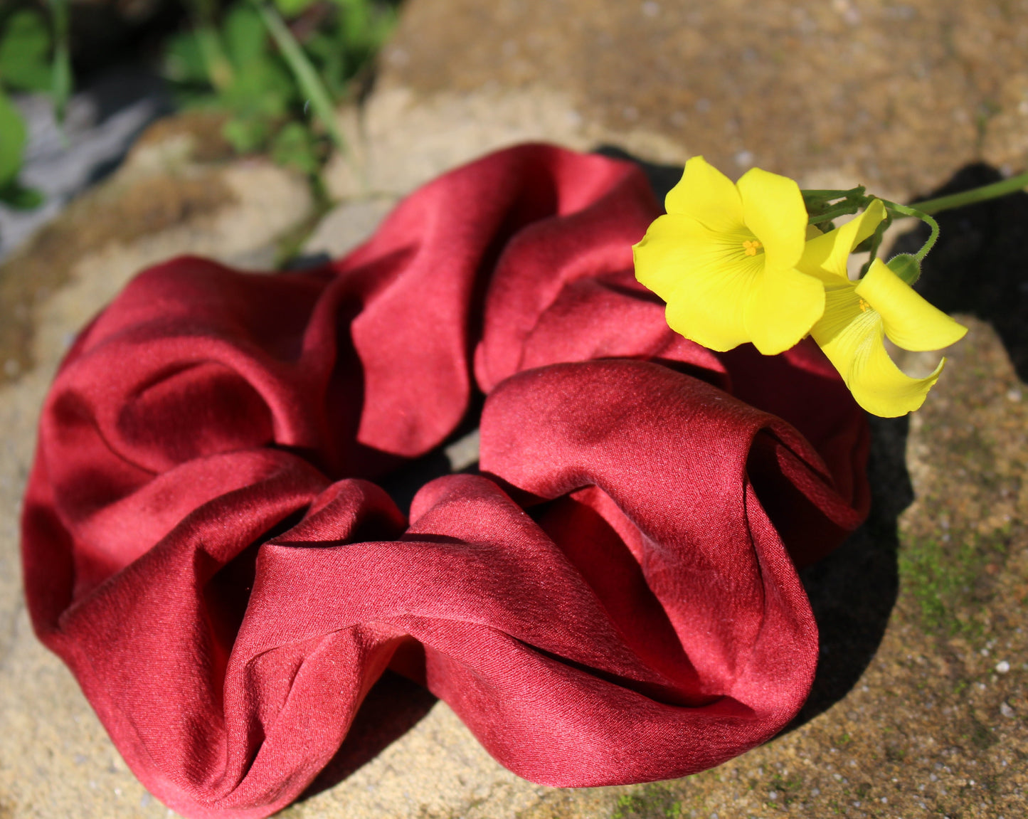 A ruby red silk scrunchie dyed with madder root to make a striking crimson red silk scrunchie, laying on a stone with a yellow flower in the frame