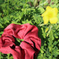 A ruby red silk scrunchie dyed with madder root to make a striking crimson red silk scrunchie, laying in a bed of clovers and a blurry yellow flower in the right corner of the frame