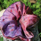 The Goose Summer St. Valentine Silk scrunchie is a plant dyed silk scrunchie with cosmic pinks and purples. one-of-a-kind silk plant dyed scrunchies. the scrunchie is lying in a bed of green clovers. 