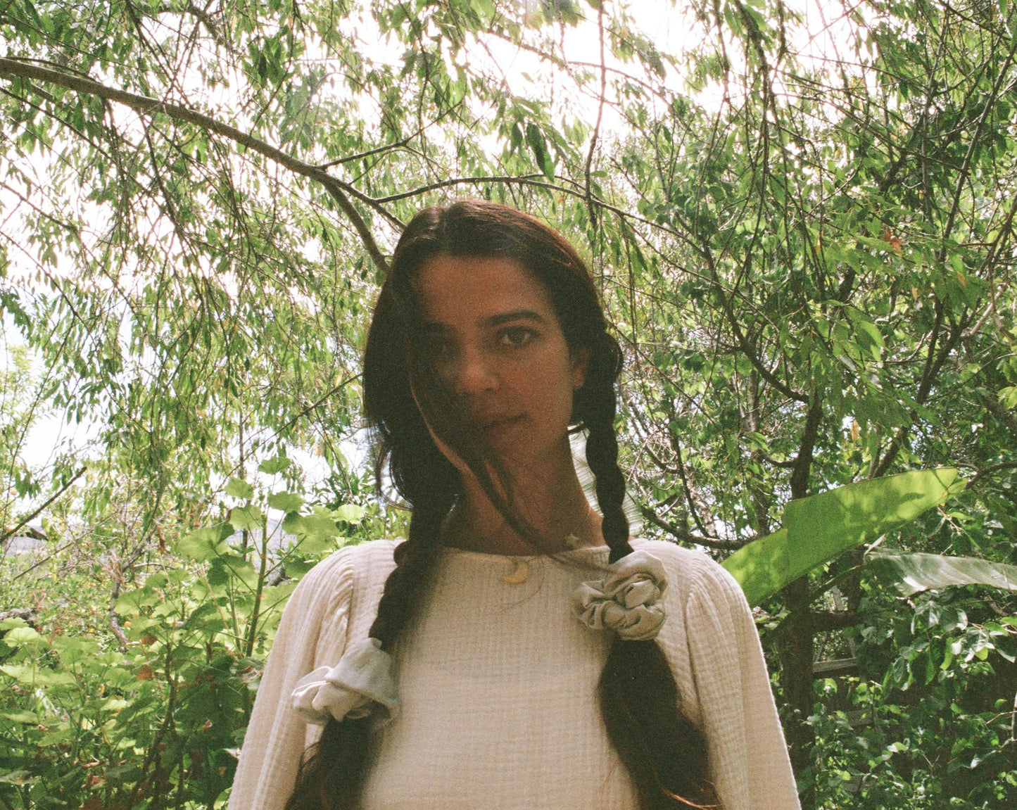 Woman wearing her hair in braids are tied with light blue naturally dyed silk scrunchies. She is wearing Goose Summer's Dawn silk scrunchies. She is surrounded by foliage. 