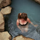 Woman swimming in a hot spring in a black bathing suit with her hair tied up in a ponytail with a Goose Summer naturally dyed brown silk scrunchie. She is wearing the Sienna Silk Scrunchie which is plant dyed with cutch bark. 