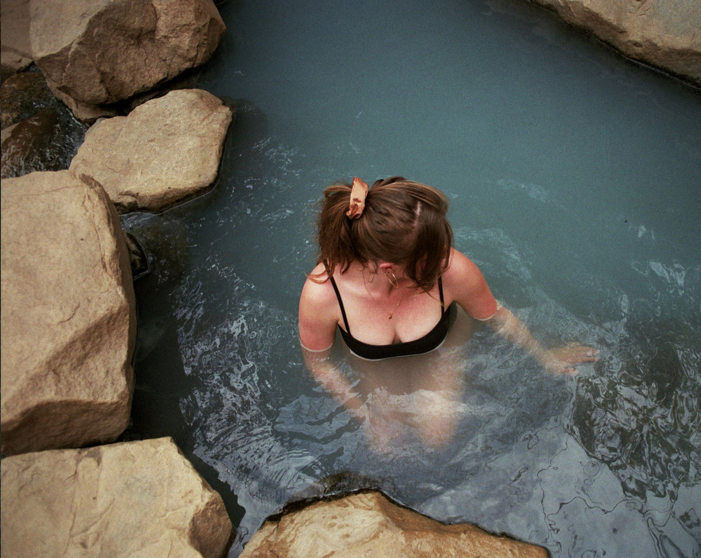 Woman swimming in a hot spring in a black bathing suit with her hair tied up in a ponytail with a Goose Summer naturally dyed brown silk scrunchie. She is wearing the Sienna Silk Scrunchie which is plant dyed with cutch bark. 