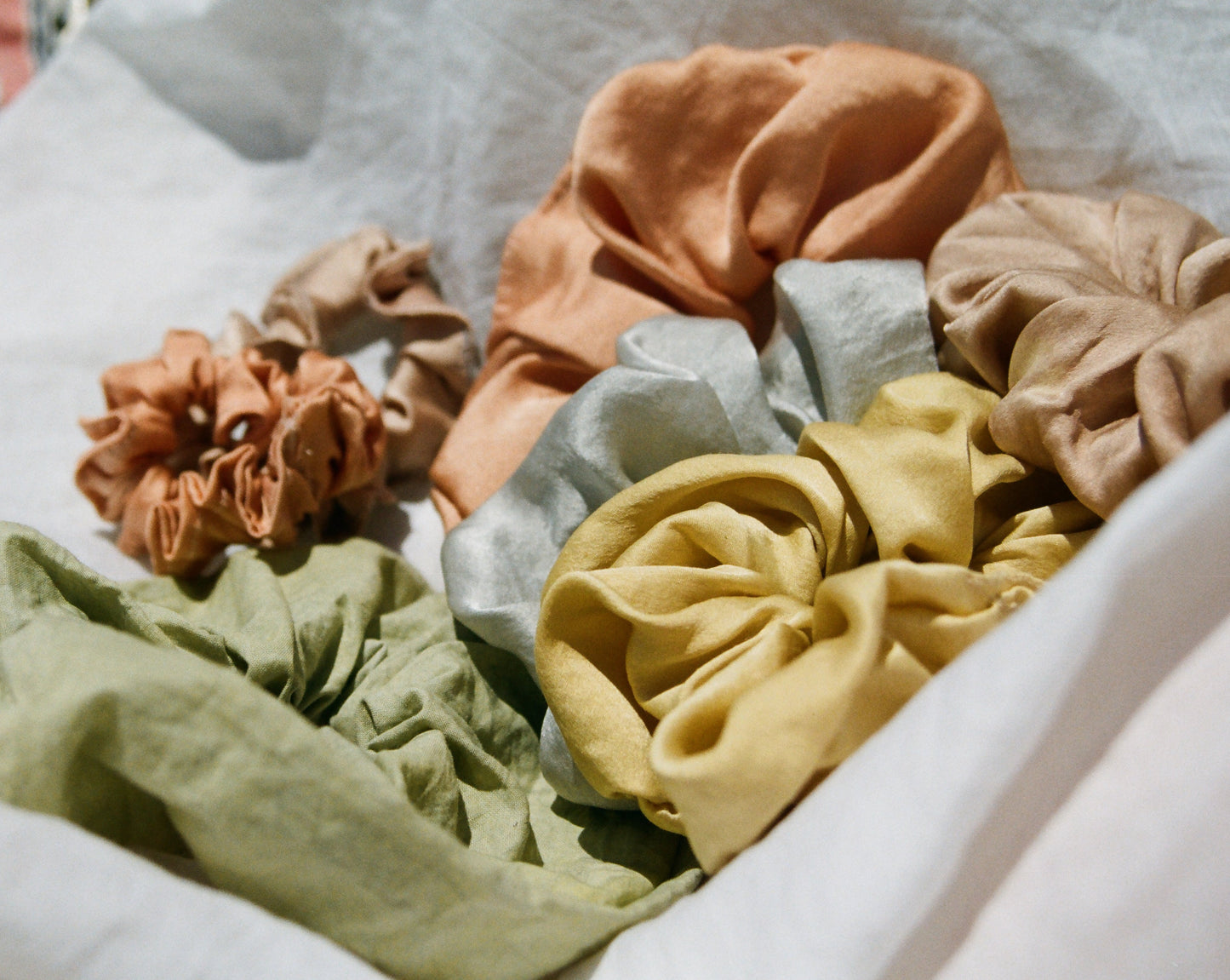 A bundle of assorted naturally dyed silk scrunchies are being displayed. There is a mint green silk scrunchie, light blue silk scrunchie, buttery yellow silk scrunchie, peach and light pink silk scrunchie. All silk scrunchies are naturally dyed.