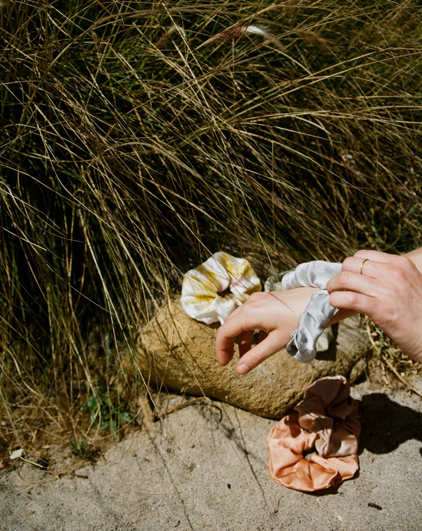 A woman is putting a light blue silk scrunchie on her hand. Behind her are more silk plant dyed scrunchies and tall grass blowing in the wind. 