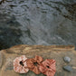 Three naturally dyed Goose Summer silk scrunchies are displayed on a rock in front of a hot spring. The colors of scrunchies from left the right are light pink, brown and rose pink silk scrunchies. Their names are Apricot scrunchie, Sienna Scrunchie, and Rose Scrunchie. 