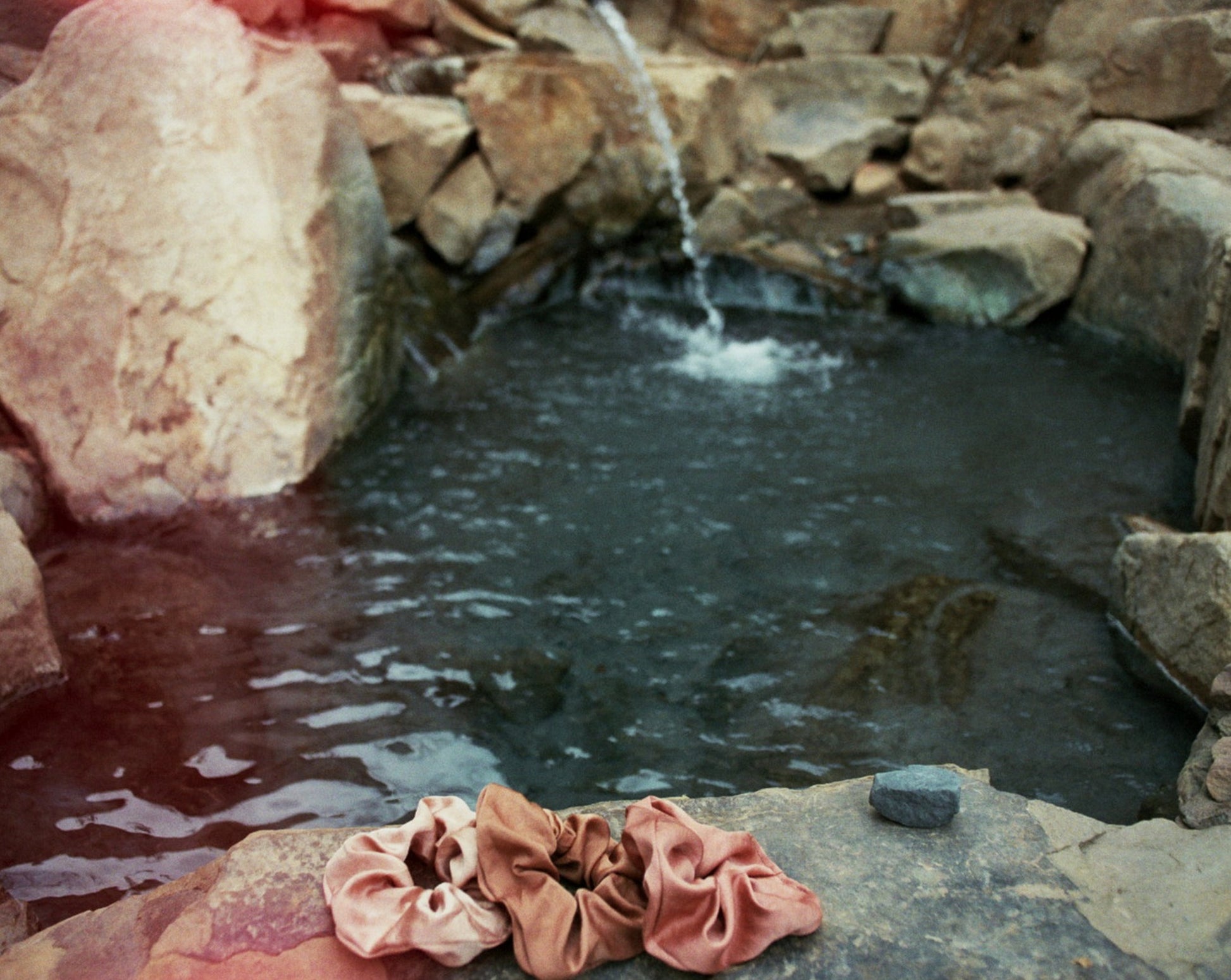 Three naturally dyed Goose Summer silk scrunchies are displayed on a rock in front of a hot spring. The colors of scrunchies from left the right are light pink, brown and rose pink silk scrunchies. Their names are Apricot scrunchie, Sienna Scrunchie, and Rose Scrunchie. 