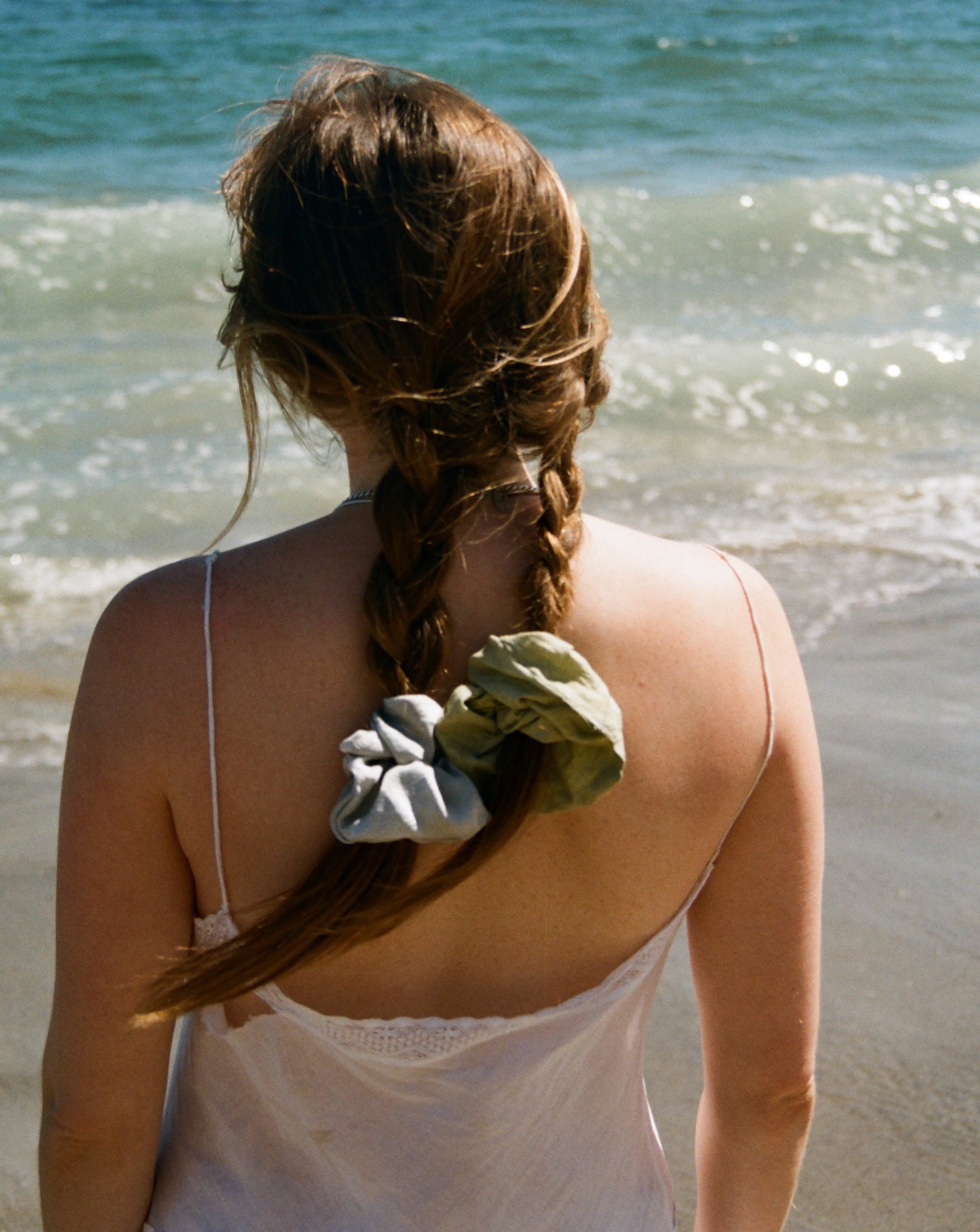 Woman standing at the beach with her back to the camera wearing her blond hair in two braids. Her braids are tied with one green and one light blue silk scrunchie. The silk scrunchies are plant dyed with indigo and weld by Goose Summer.