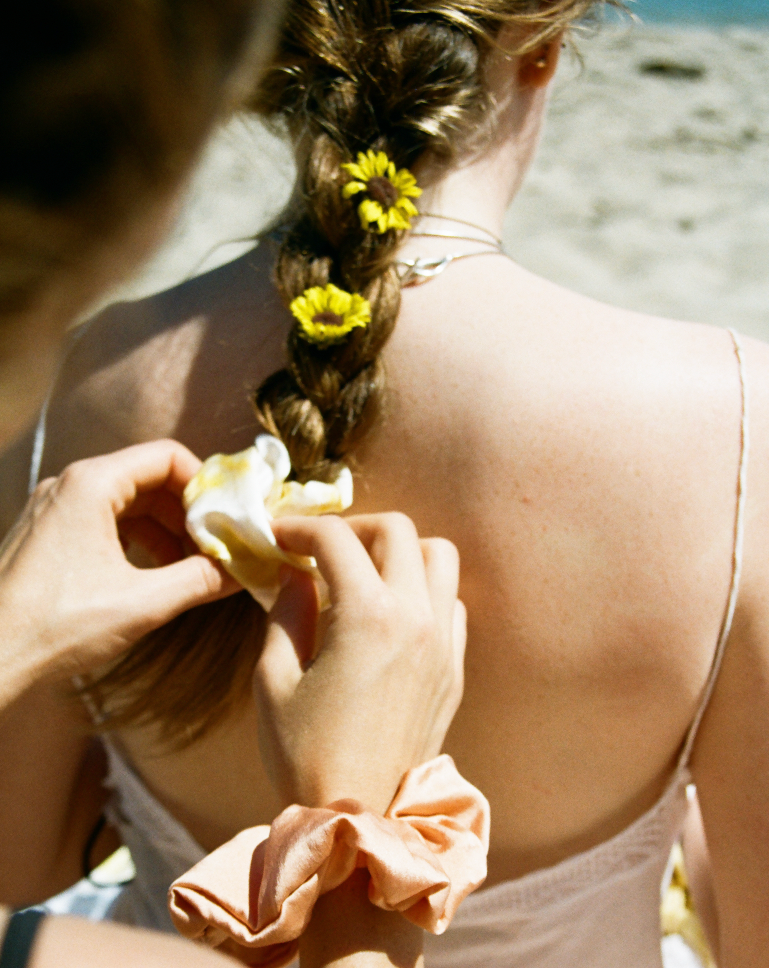 Two woman sitting on the beach, one woman is tying another woman's hair into a braid and using yellow flowers and a Goose Summer naturally dyed silk scrunchie in her hair. She also has a peach silk scrunchie on her wrist. 