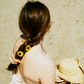 Woman sitting in the sand on the beach wearing her hair in one braid tied with a peach silk scrunchie. Peach is dyed naturally with madder root by Goose Summer. Madder Root is a plant dye that has been cultivated for hundreds of years as a natural textile dye. She has a peach scrunchie and yellow wild flowers in her hair. 