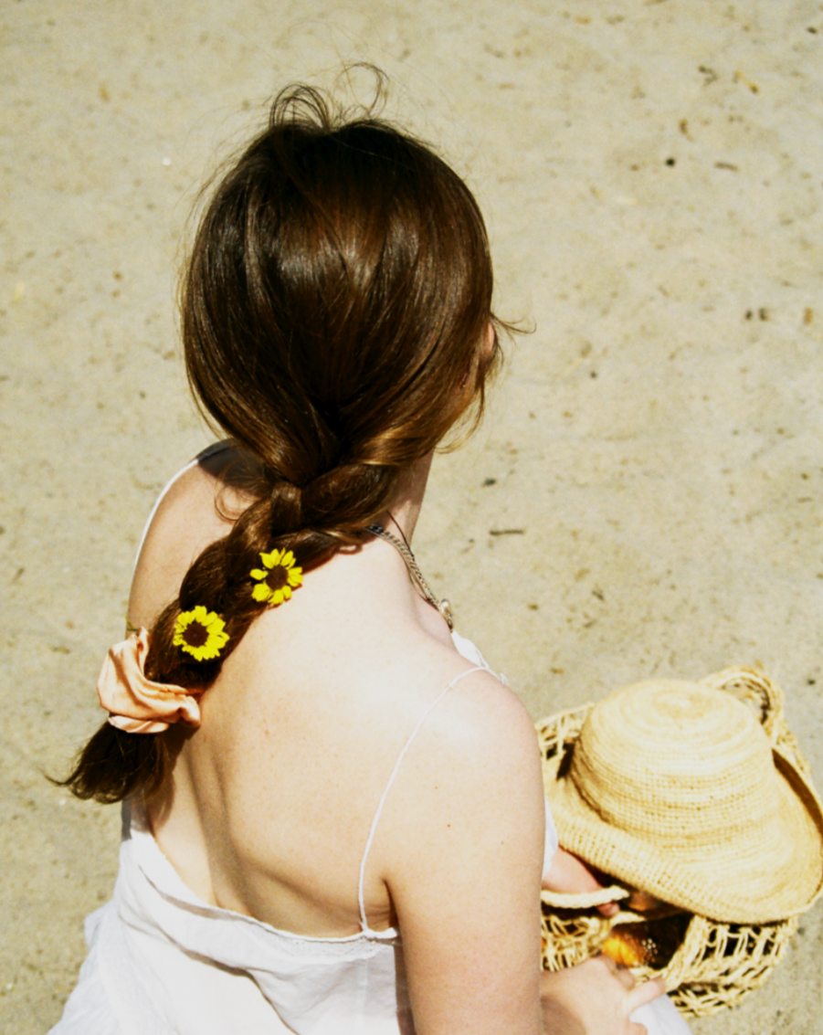 Woman sitting in the sand on the beach wearing her hair in one braid tied with a peach silk scrunchie. Peach is dyed naturally with madder root by Goose Summer. Madder Root is a plant dye that has been cultivated for hundreds of years as a natural textile dye. She has a peach scrunchie and yellow wild flowers in her hair. 